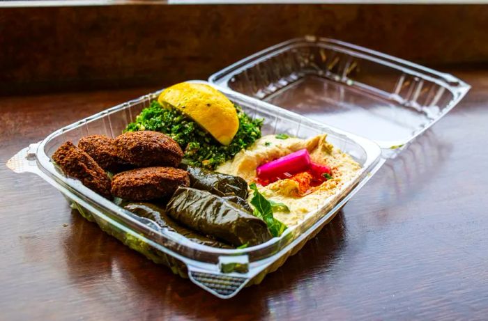 A plastic takeout container brimming with stuffed grape leaves, hummus, falafel, and tabbouleh.