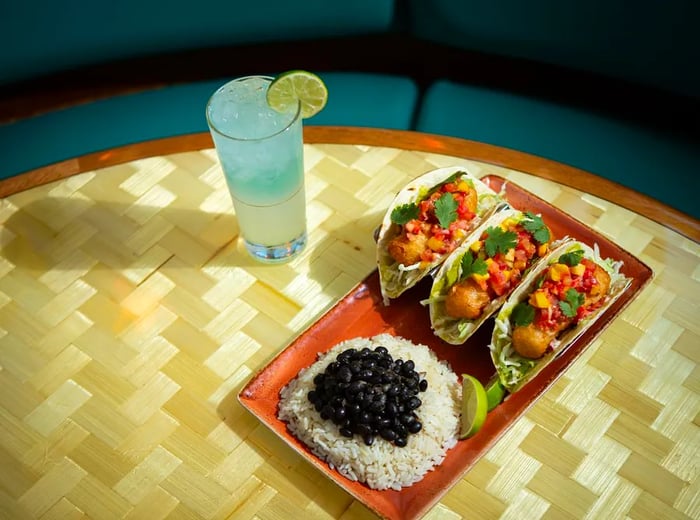 A blue cocktail rests on a table alongside three fish tacos accompanied by rice and beans.