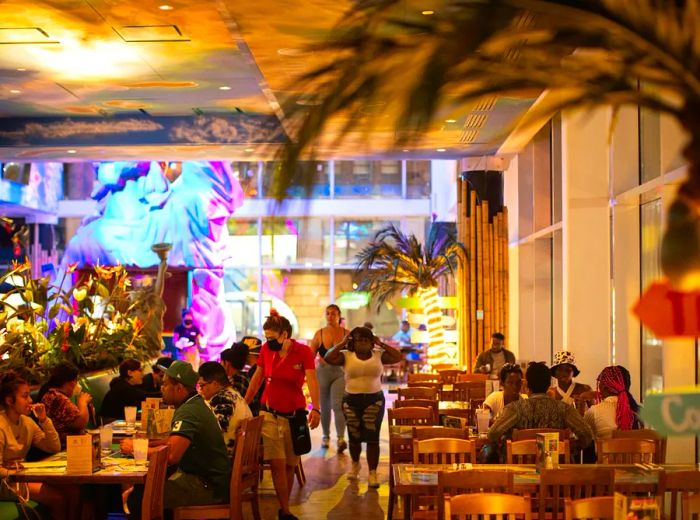 Tourists enjoy their meals at wooden tables within a tropical-themed dining room.