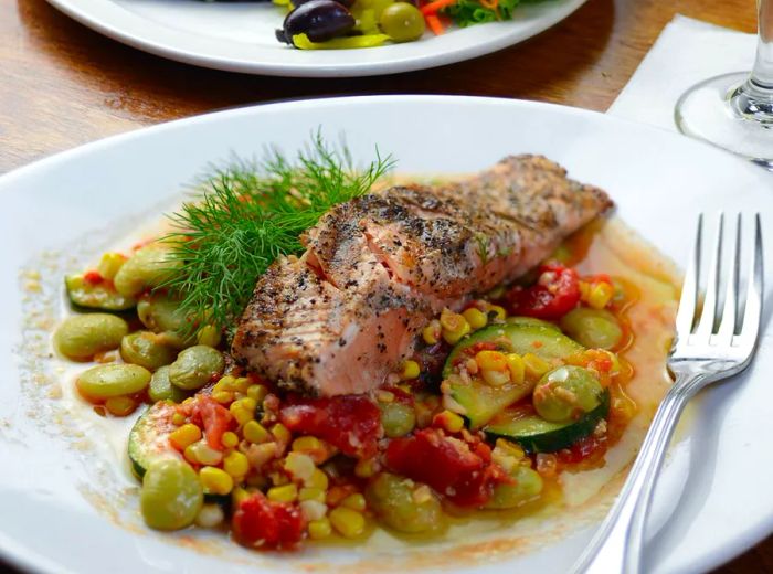 A perfectly grilled salmon fillet accompanied by sweet potatoes is served over a vibrant succotash featuring a medley of summer vegetables and lima beans, all topped with a fresh sprig of dill.