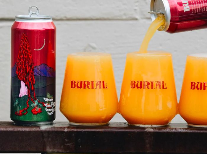 A hand is pouring a beer from a can into three glasses, causing them to overflow over the railing they rest on, next to another full can featuring a decorative label.