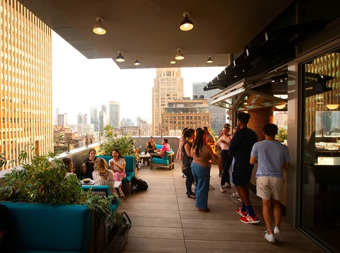 A lively rooftop restaurant scene, with people chatting around the bar and seated in booths, set against a city backdrop.