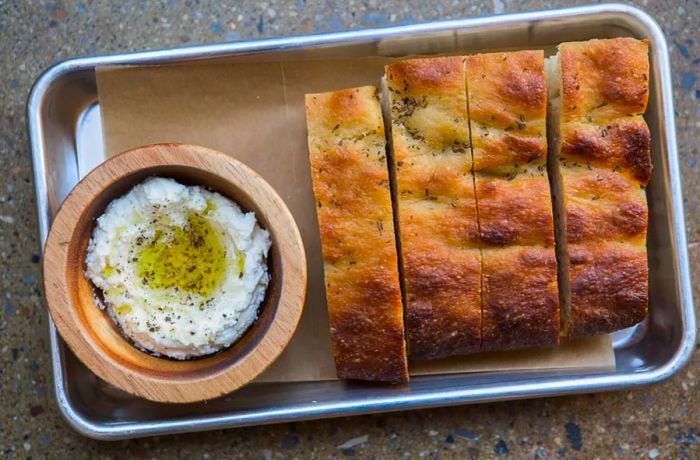 A metal tray featuring thick slices of bread alongside a wooden bowl of ricotta drizzled with olive oil.