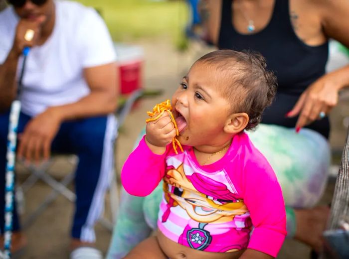 a baby in a pink swimsuit clutches a handful of spaghetti to her mouth.