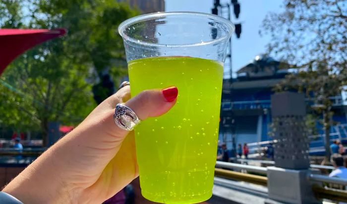 A hand lifts a clear plastic cup filled with a yellow, bubbly drink.