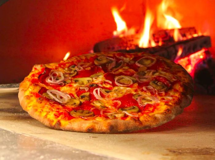 A complete pizza pie, angled to showcase its toppings, nestled in a wood-fired oven. The pizza is adorned with sliced mushrooms and onions.