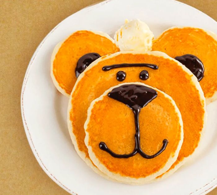 A stack of pancakes in various sizes, creatively decorated with chocolate syrup to resemble a bear's face.