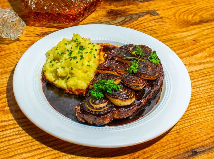 A serving of steak with creamy mashed potatoes, drizzled in sauce, placed on a wooden surface next to a floral pot.