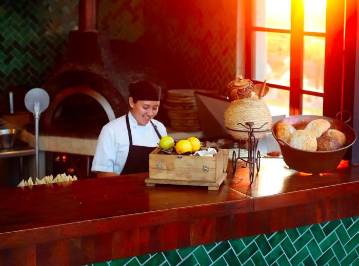 A chef wearing an apron works in an open kitchen with a wood-fired oven in the background.