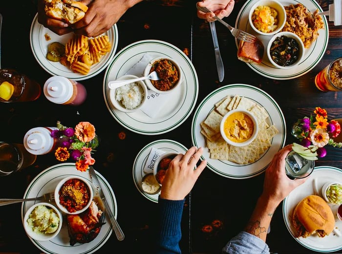 An aerial view of a table overflowing with barbecue delights. Several hands reach for sandwiches, pulled pork, mac and cheese, beans, BBQ chicken, and more.