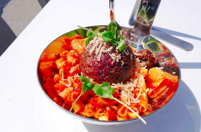 A meatball rests atop a bed of pasta in a large metal spoon.