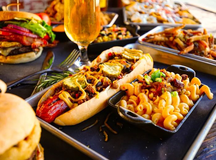 A platter showcasing a large hot dog topped with chili and cheese, accompanied by a cast iron dish of mac and cheese, along with various burgers, sandwiches, and beverages on a wooden table.