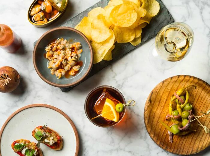 An aerial view of a selection of tapas and snacks—canned mussels, potato chips, olives, and more—arranged on a marble countertop next to a glass of white wine.