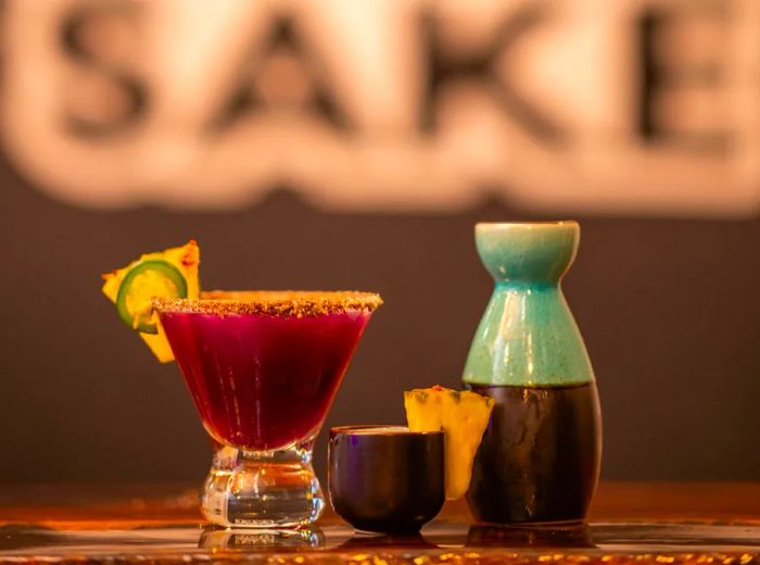 A vibrant carafe alongside a bright purple cocktail and a small sake cup adorned with a pineapple wedge, resting on a wooden surface.