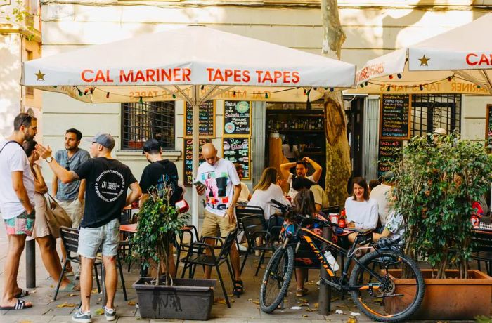 Outside a bustling restaurant, patrons are gathered under umbrellas, some seated while others stand, alongside parked bicycles.