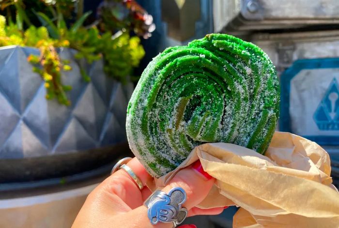 A hand presents a green swirled pastry wrapped in paper.