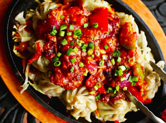 From above, a plate of dumplings adorned with a vibrant sauce and sprinkled with chopped chives, with metal tongs resting on the side.