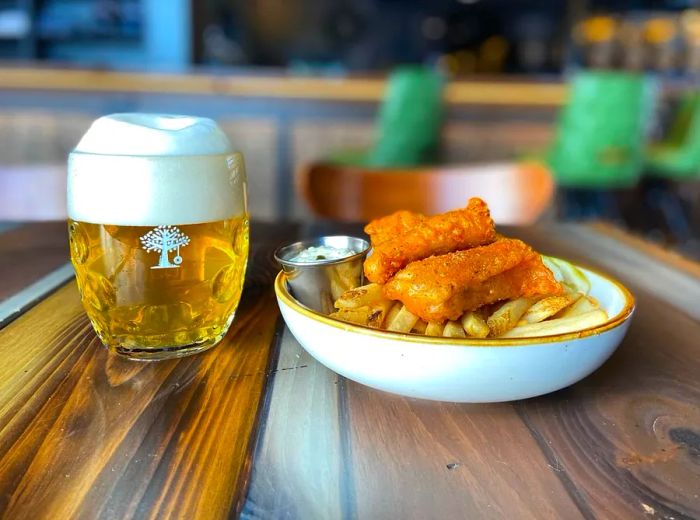 A plate featuring crispy fried fish atop a bed of french fries, paired with a pint of beer on a rustic wooden table.