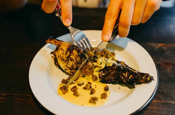 Hands skillfully cut into a charred wedge of cabbage drizzled with a yellowish sauce using a knife and fork.