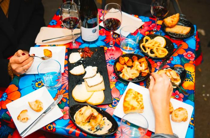 A vibrant tablecloth displays an array of small dishes filled with cheese, potatoes, and wine.