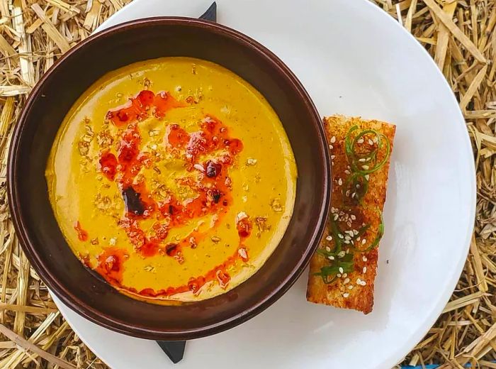 An overhead view of a bowl filled with vibrant red soup, accompanied by a slice of garlic toast on a plate.