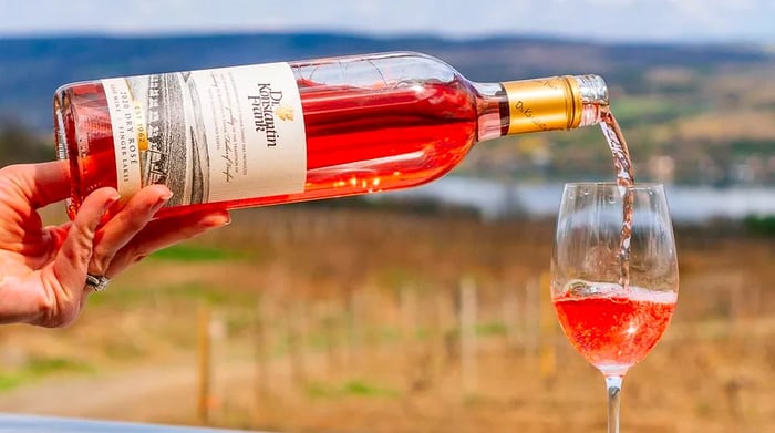 A hand pours rosé into a wine glass, set against a backdrop of a sunny vineyard in spring.