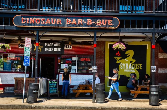 A red-brick restaurant façade features a server carrying a tray between outdoor picnic tables, with a sign that reads Dinosaur Barbecue.