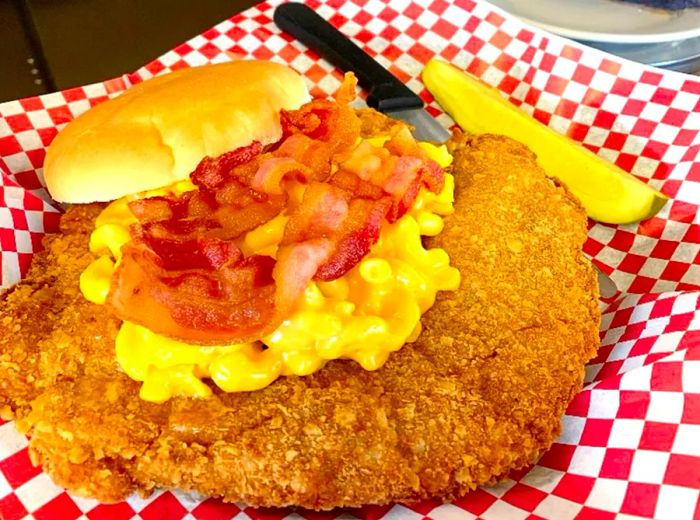 An enormous fried pork tenderloin lavishly topped with mac and cheese, crispy bacon, and a bun, presented in a basket lined with checkerboard paper.