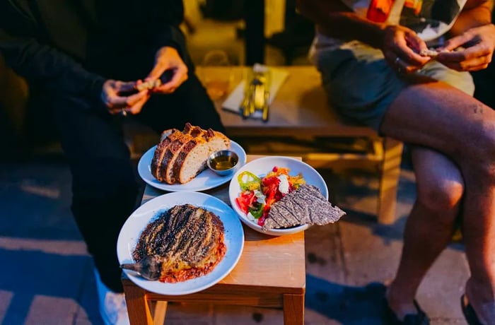 On a small sidewalk table, three modern dishes are displayed while two customers are seen in the background, ready to enjoy their meals.