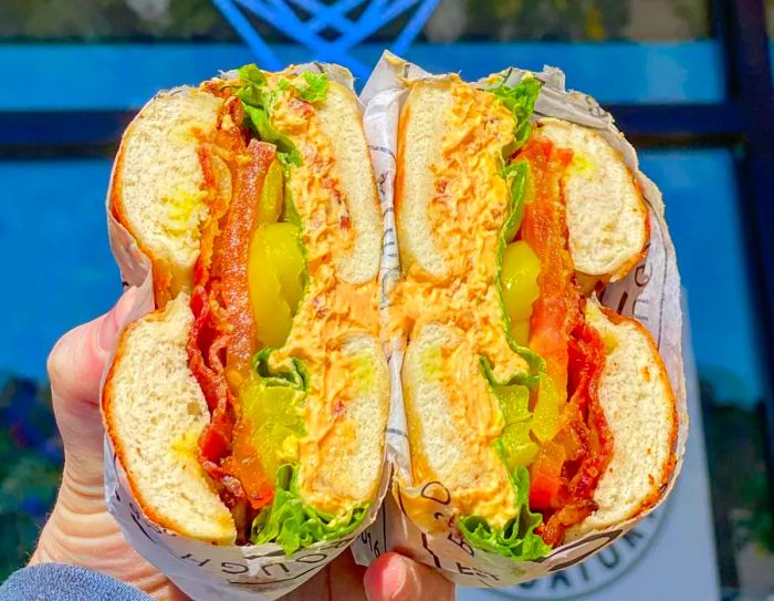 A bagel sandwich sliced in half showcases layers of meat, fresh vegetables, and flavored cream cheese, positioned in front of a glowing ‘open’ sign.