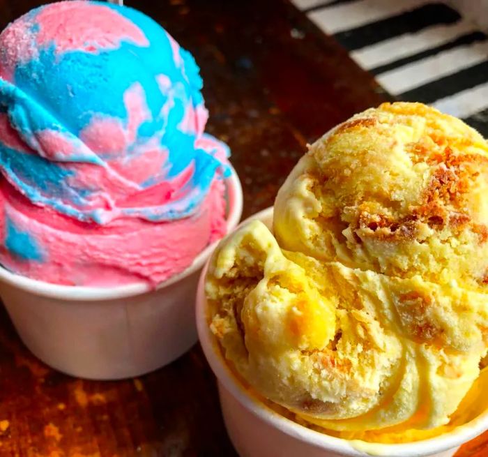 Two ice cream cups: one featuring a vibrant blue and pink swirl, the other a creamy buttery flavor dotted with pieces of Twinkie.