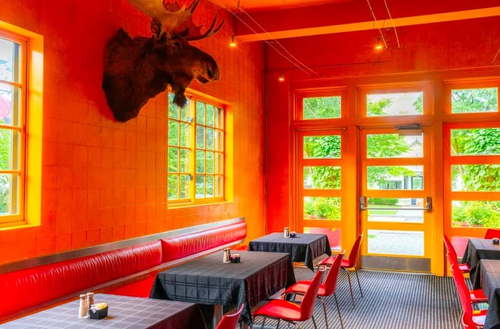 A bright, wood-paneled restaurant interior featuring a large moose head as decor, red leather banquettes, and lush greenery visible through expansive windows.