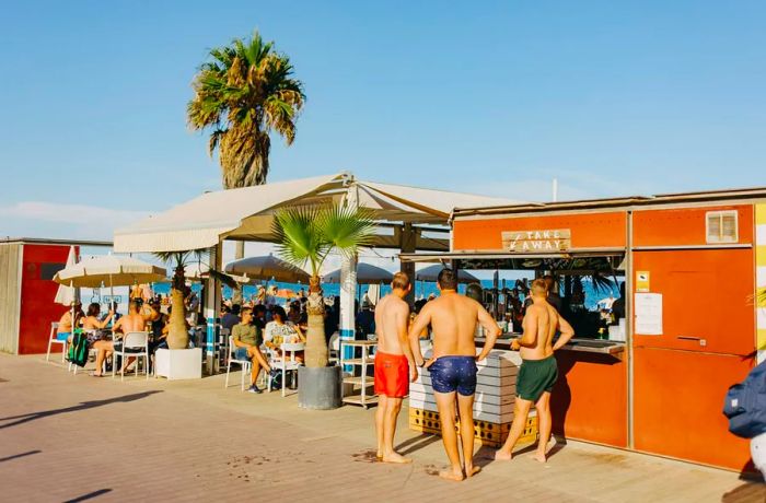 Three men in swim trunks gather at a takeaway window along a sunlit boardwalk.