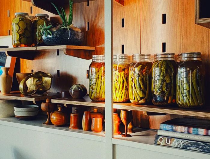 Shelves adorned with jars of vibrant vegetable pickles sit beside wooden serveware, artwork, and cookbooks.