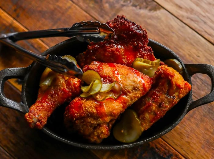 A cast iron skillet brimming with half a fried chicken garnished with pickles, tongs resting on the skillet's edge atop a wooden table.