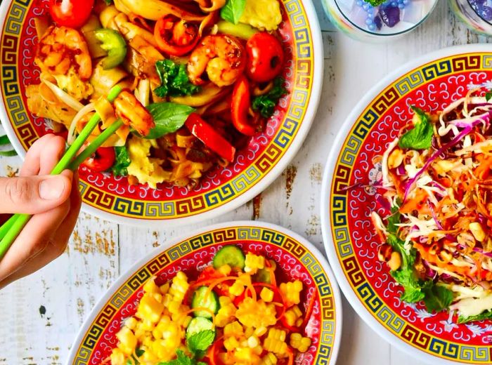 A table spread with a variety of dishes, featuring a plate of stir-fried noodles that someone is enjoying with chopsticks, alongside banana blossom salad and a corn salad.