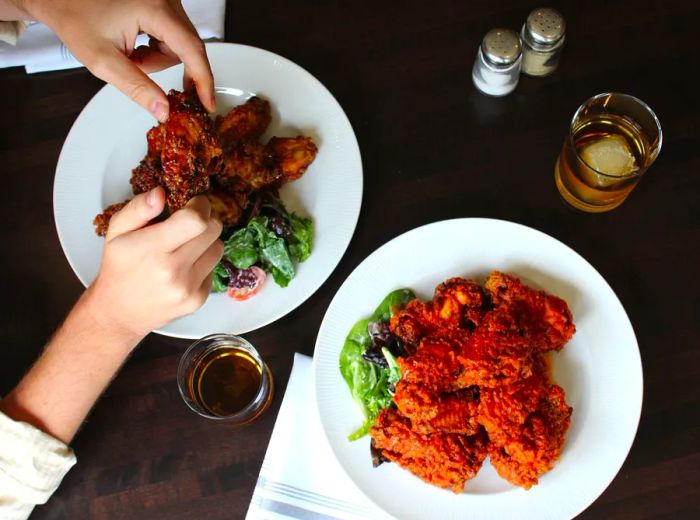 A hand reaches for a crispy piece of fried chicken from a plate, accompanied by another plate and a glass of whiskey nearby.