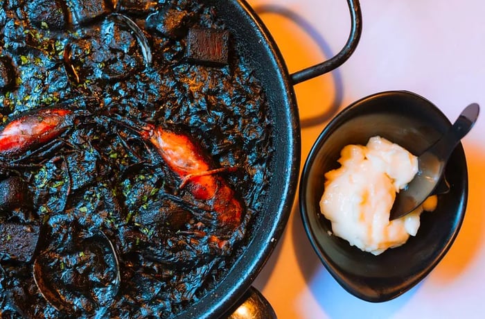 A round dish of black rice accompanied by a side of white aioli.