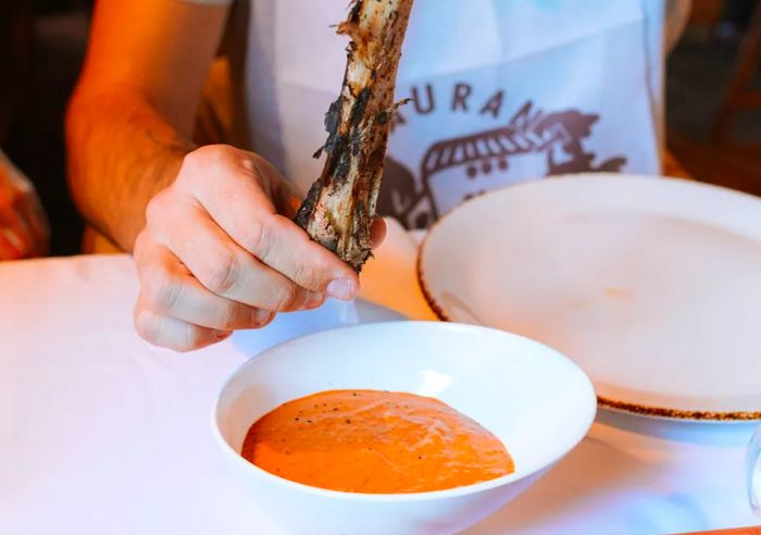 A hand holds a charred green onion above a bowl of vibrant orange romesco sauce.