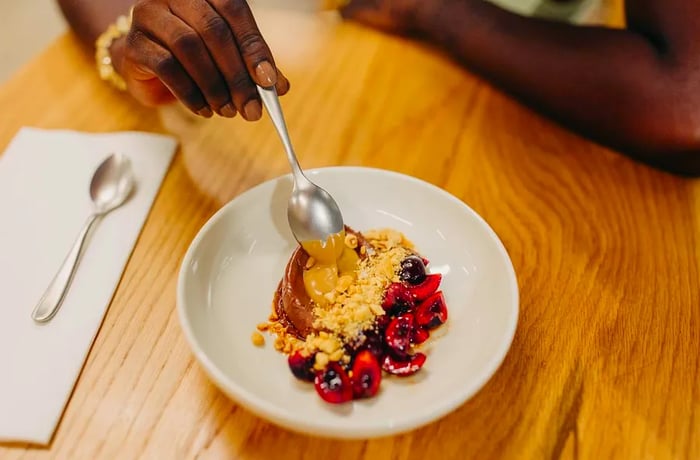 A hand reaches for a spoon, ready to enjoy a dessert featuring fresh cherries and delightful accompaniments on a wooden table.