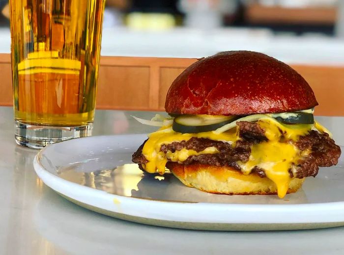 A towering double burger loaded with gooey melted cheese and pickles sits on a plate next to a pint of beer.