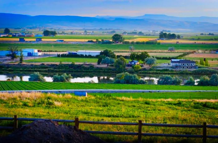 A picturesque rural landscape featuring farms nestled around a river, framed by an expansive sky and distant mountains.