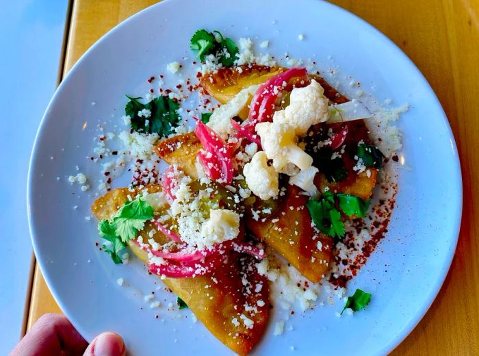 A hand presents a plate of empanadas adorned with cauliflower florets, crumbled cheese, onion slices, and fresh herbs.