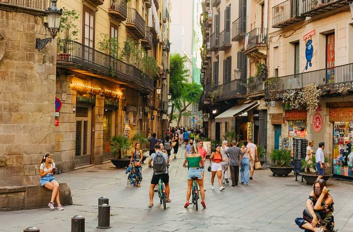 A man and woman on bicycles navigate a bustling but not overcrowded pedestrian street flanked by tall historic buildings and local shops at street level.