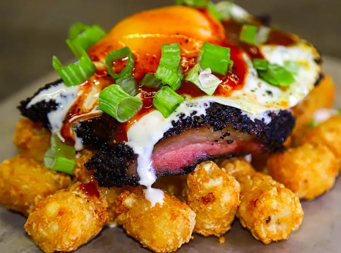 A mountain of tater tots crowned with thick slices of smoky meat, a fried egg, savory sauce, and fresh herbs.