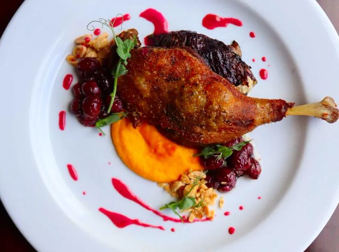 An aerial view of a plate showcasing a duck leg resting atop a vibrant orange puree, elegantly garnished with various accompaniments.