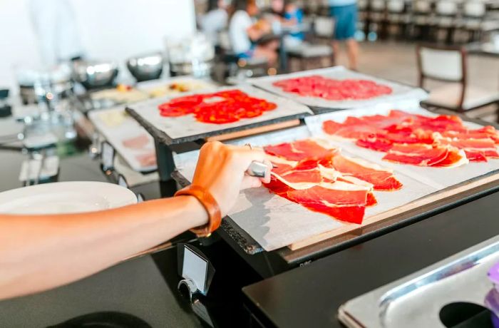 A hand reaches into the frame to grab a slice of jamón from a stacked pile.