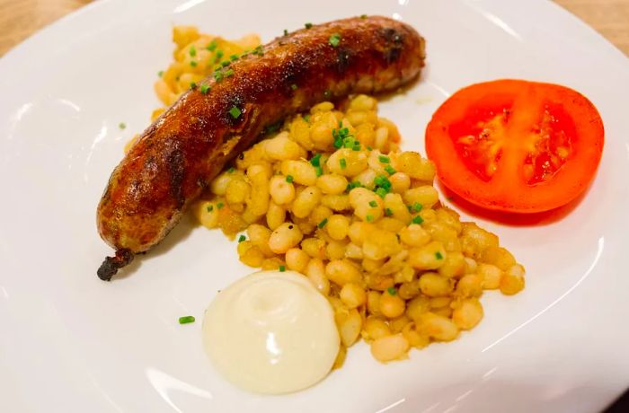 A sausage served on a plate alongside white beans, a cooked tomato, and a dollop of white sauce.