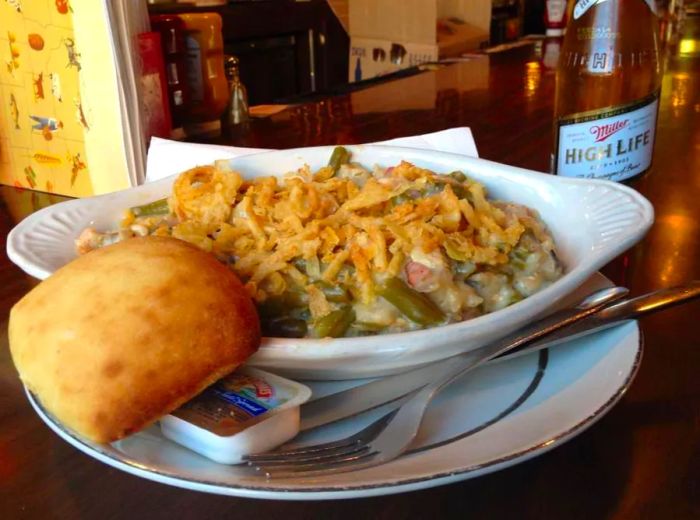 A serving dish holds a green bean casserole topped with crispy fried onions, accompanied by a roll and a bottle of High Life on a wooden bar.