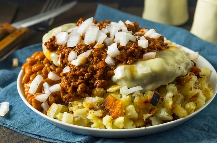 A generous serving of fried potatoes, macaroni salad, and baked beans, all topped with a cheese-covered hamburger patty, chili, and onions